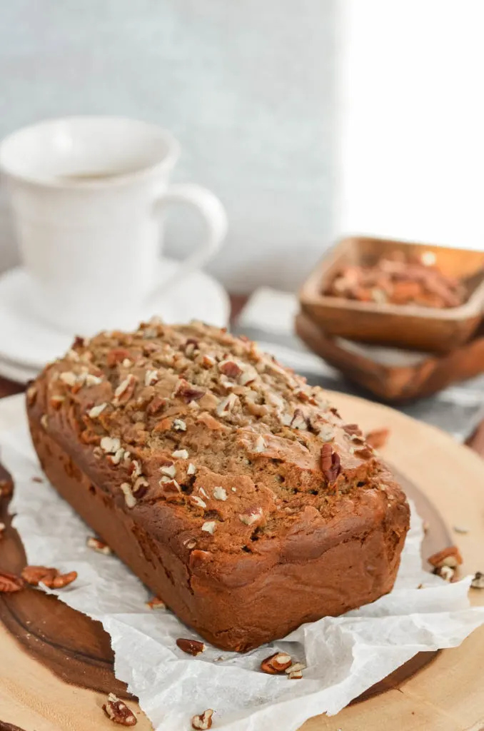 Sour Cream Banana Bread with Pecans on a wooden cutting board. A small bowl of pecans and white plated are slightly out of focus in the background. 