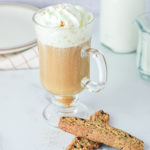 Clear Glass Coffee Mug filled with hot coffee lightened with Homemade Sweet Cream Coffee Creamer, a brown and white napkin is in the background slightly out of focus, along with a small pitcher of coffee creamer. 2 cookies sit beside the cup of coffee on a white counter top.