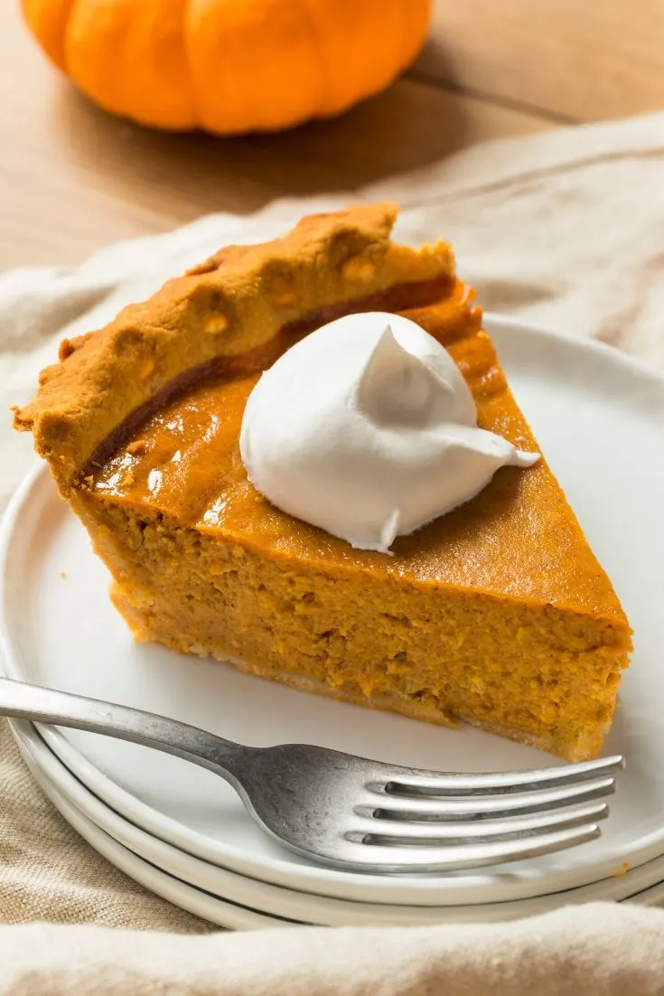 A slice of pumpkin pie sitting on a stack of 3 white plates with a fork. There's a linen napkin on the wooden table and a small pumpkin in the background. 