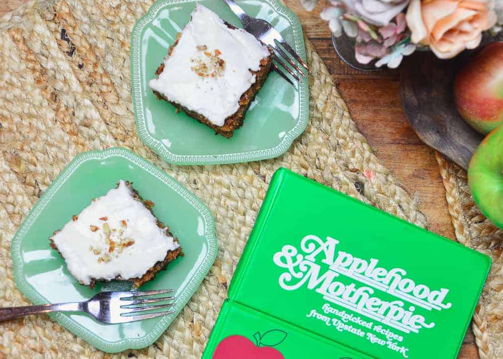 Two slices of Apple Spice cake on jade plates with forks on a wooden table sitting next to a bowl of apples and the Applehood and Motherpie Cookbook