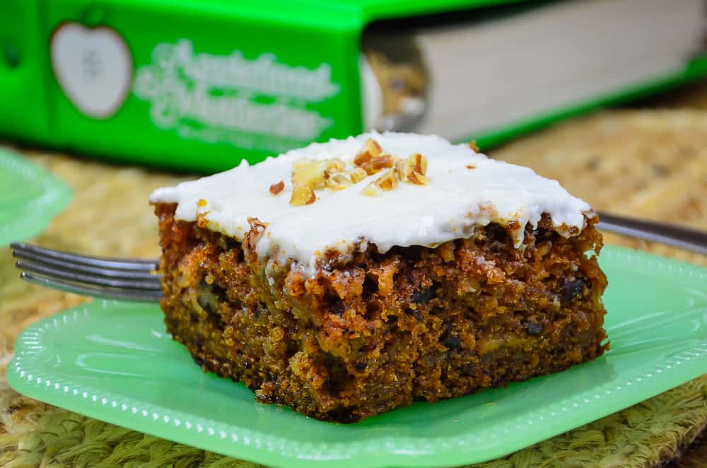 A slice of apple spice cake with cream cheese frosting on a jade plate sitting in front of the cookbook Applehood and Motherpie