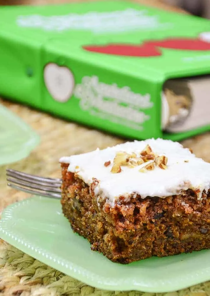 A slice of apple spice cake with cream cheese frosting on a jade plate sitting in front of the cookbook Applehood and Motherpie