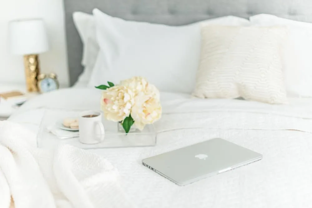 Macbook on bed with a tray holding cookies and coffee. Alarm clock and lamp on bedside table.