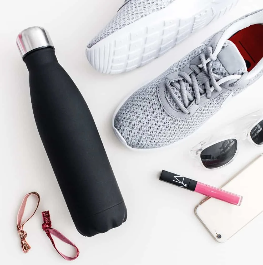 Water bottle, lip gloss, hair ties, sneakers and sun glasses laid out on a white surface