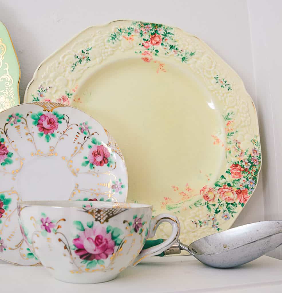 Large vintage dinnerplate with floral pattern on a white shelf, with a withe teacup with a rose pattern and vintage measuing scoop. 