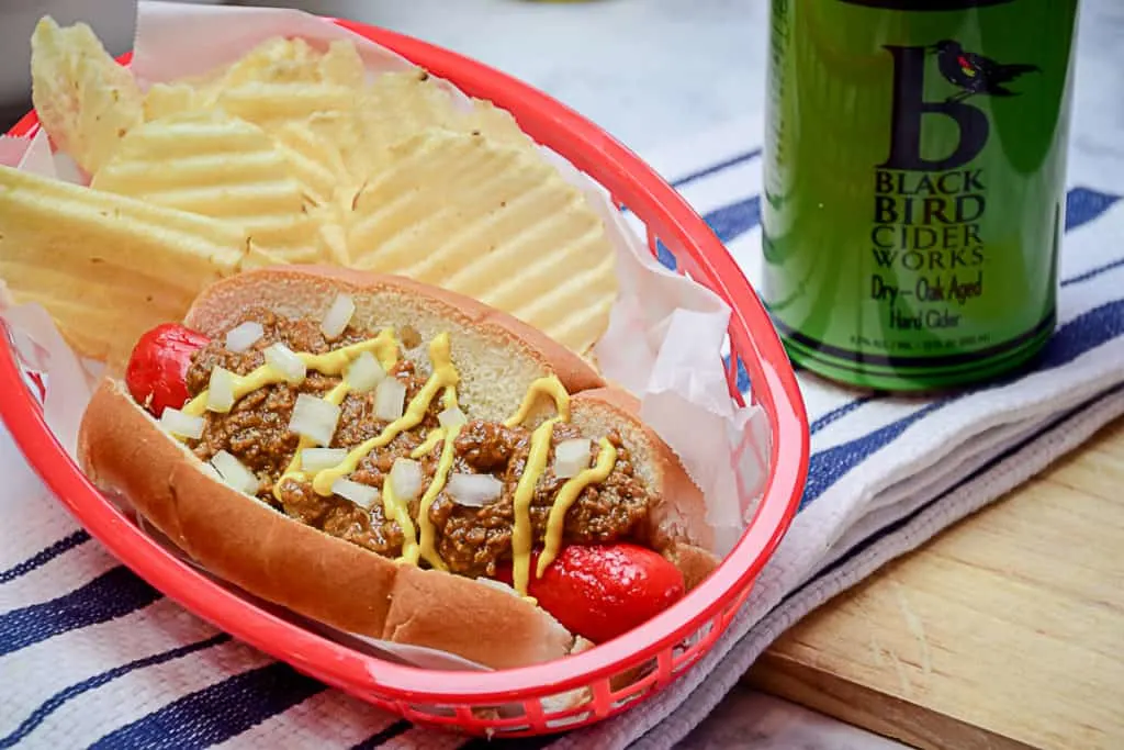 A red plastic food basket holding a Michigan style hot dog with mustard and chopped onions on a new england style bun, sitting next to a can of Upstate New York hard cider. 