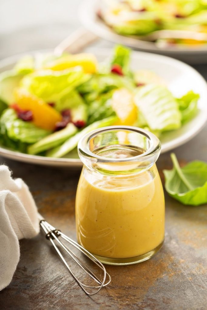 A small glass jar of golden homemade salad dressing on a table  sitting next to a whisk and napkin, with a salad slighlty out of focus in the background. 