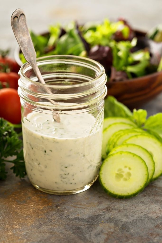A mason jar full of a creamy  homemade salad dressing with a serving spoon in it on a table surrounded by fresh cucumbers, tomatoes and lettuce, with a bowl of salad slightly out of focus in the background. 
