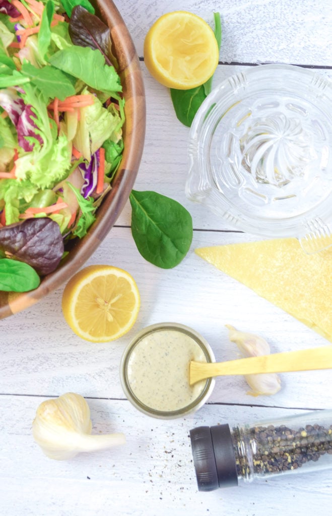 Homemade Garlic Pepper Parmesan Salad Dressing in a jar surrounded by lemons, a wedge of parmesan cheese, a big bowl of salad greens, and a pepper grinder. 