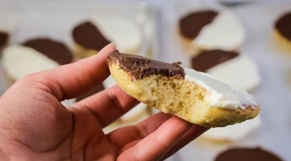 The side view of a hand holding a vanilla half moon cookie that has been bit into, with frosted half moon cookies on cookie sheets slightly out of focus in the background. 