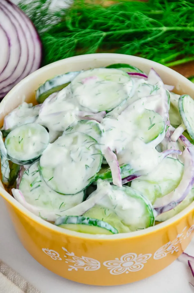 Creamy Cucumber Salad with red onion in a vintage yellow Pyrex mixing bowl on a marble countertop. A wooden cutting board with fresh dill and sliced cucumbers are slightly out of focus in the background. 