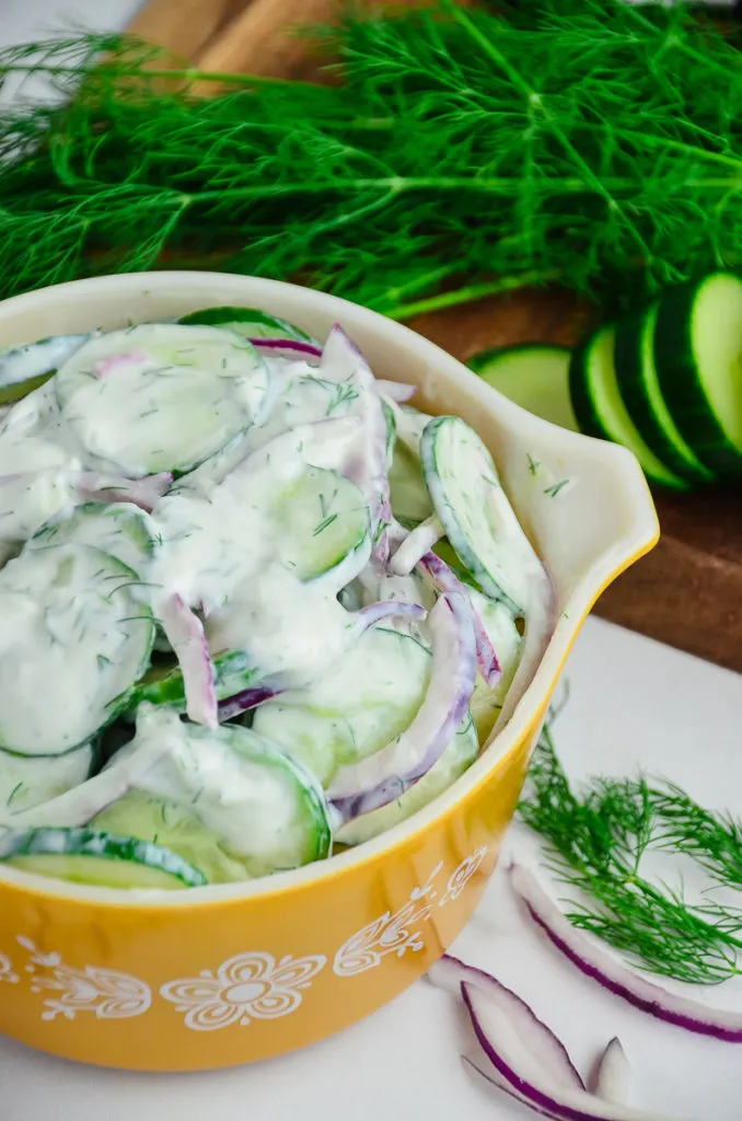 Creamy Cucumber Salad with red onion in a vintage yellow Pyrex mixing bowl on a marble countertop. A wooden cutting board with fresh dill and sliced cucumbers are slightly out of focus in the background. 