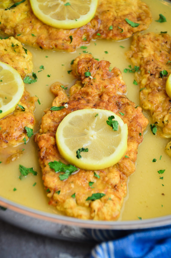 Close up of chicken french, with a lemon slice on top of a golden brown chicken cutlet, in a lemon wine pan sauce sprinkled with chopped parsley. 