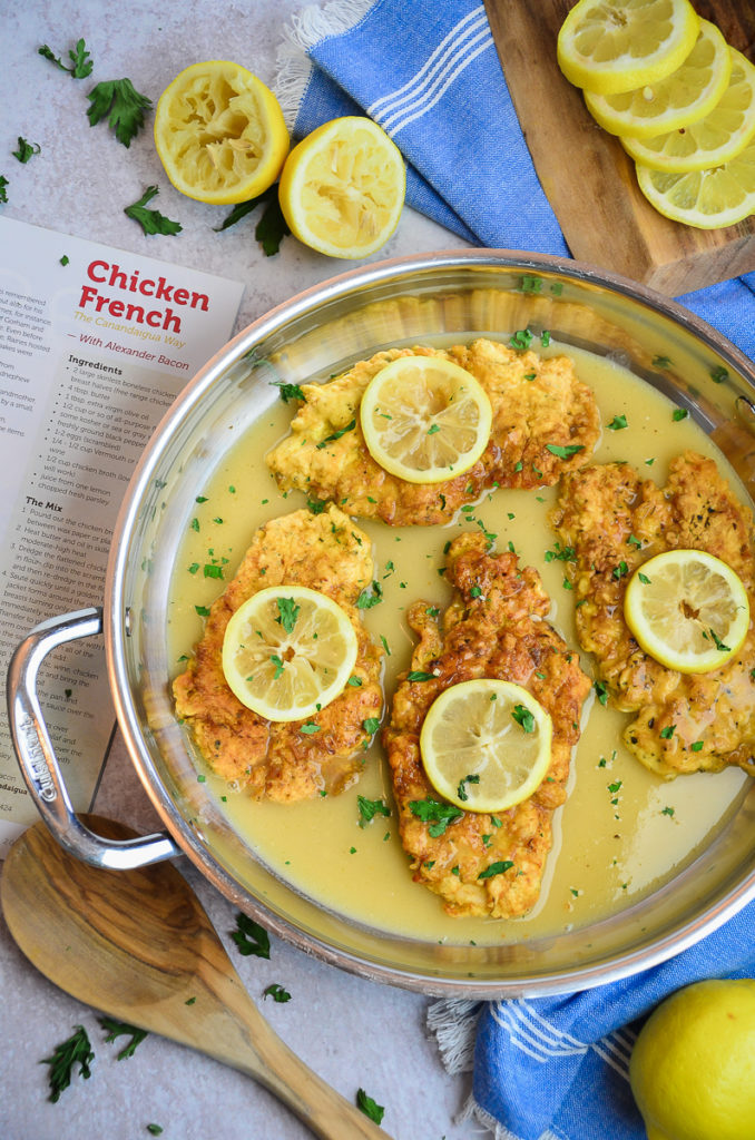 A skillet with 4 servings of chicken french, each cultlet has a lemon slice on top of a golden brown chicken breast, in a lemon wine pan sauce sprinkled with chopped parsley. 