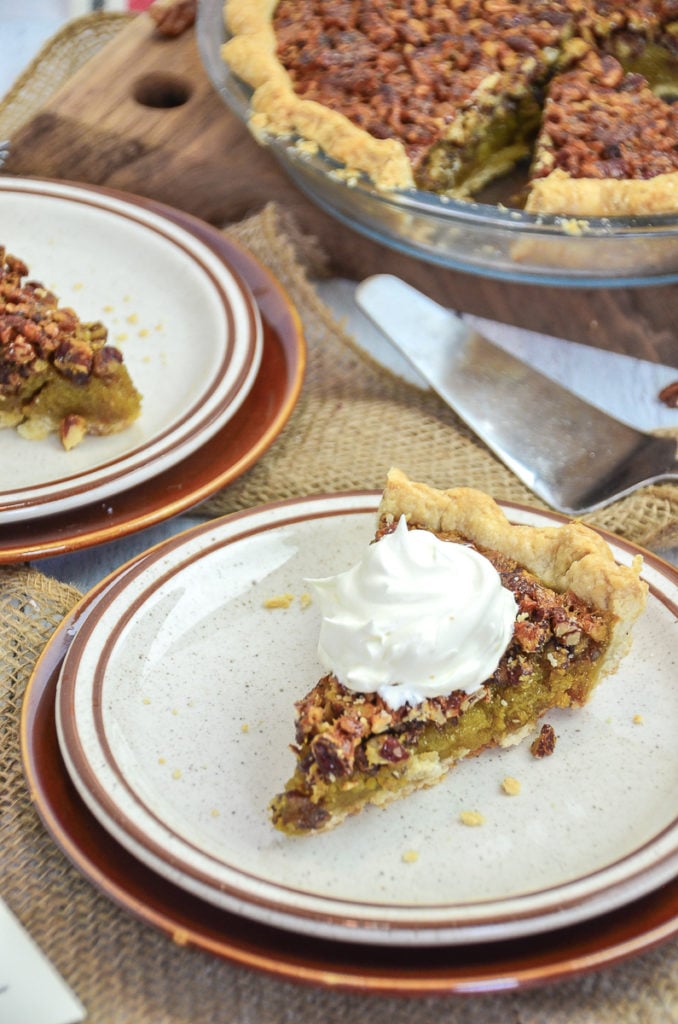 A close up view of two brown and white plates that each have 1 slice of pecan pie, with the remainder of the pie in the pie dish sitting slightly out of focus. 1 of the slices of pecan pie is topped with fresh whipped cream.