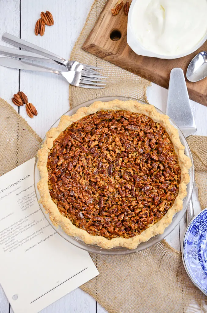 Overhead view of homemade pecan pie on a white table. The recipe for the Kopper Kettle pecan pie is sitting beside the pie. 