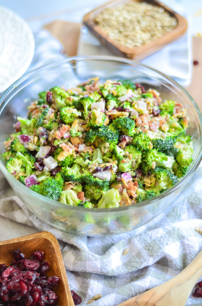 A large bowl of fresh broccoli salad on a wooden table . A stack of plates are slightly out of focus in the backgroun