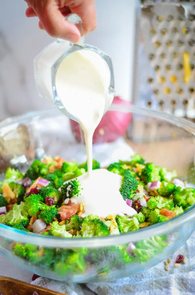 Broccoli salad in a large clear glass bowl. A creamy maoy-based dressing is being poured onto the salad from a small glass pitcher. 