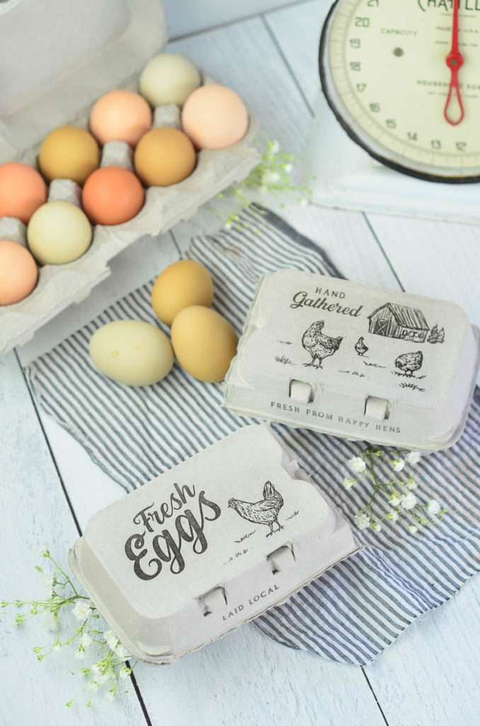 2 half dozen close egg cartons sitting on a white table tops. One carton is marked "Hand Gathered" and has a banyard scene with chickens on it, the other is marked with "Fresh Eggs" with a chicken graphic on it.  there are a dozen multi colored fresh eggs slightly out of focus in the background and a vintage kitchen scale. 