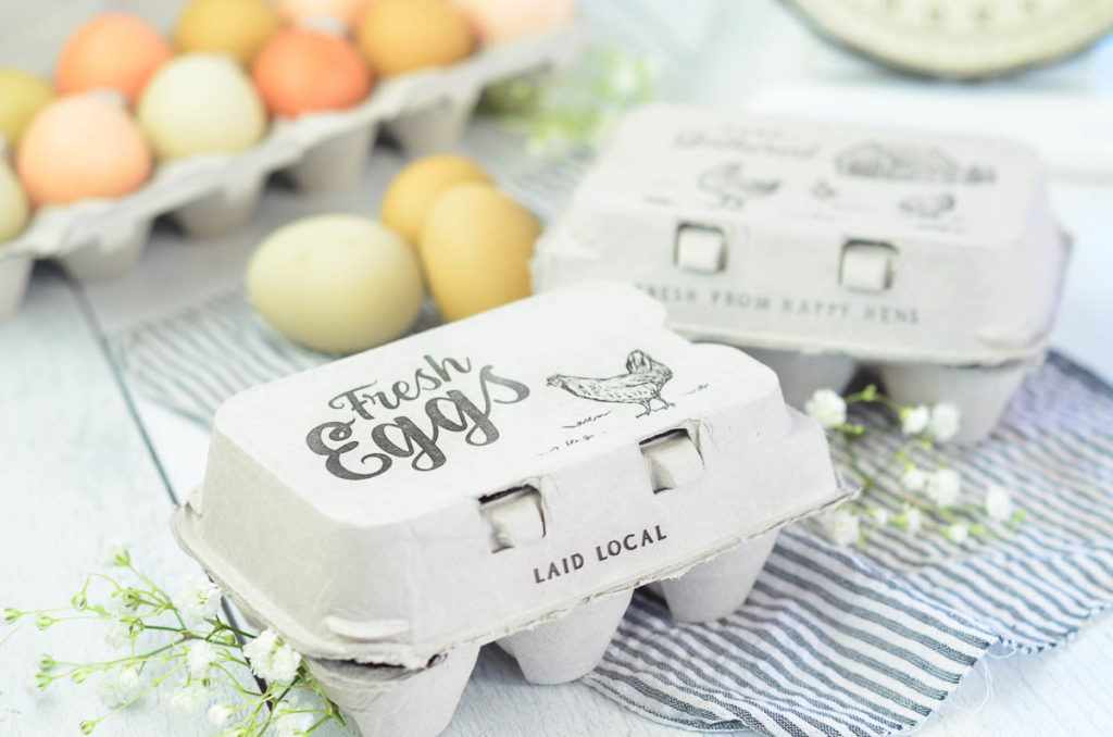2 half dozen close egg cartons sitting on a white table tops. One carton is marked "Hand Gathered" and has a banyard scene with chickens on it, the other is marked with "Fresh Eggs" with a chicken graphic on it. there are a dozen multi colored fresh eggs slightly out of focus in the background and a vintage kitchen scale. 