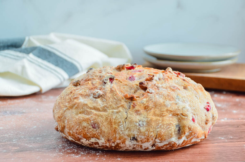 An whole loaf of cranberry pecan no-knead bread on a wooden table, with plates and a kitchen towel slightly out of focus in the background. 
