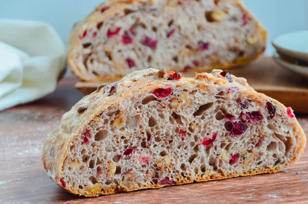 Cross section view of a cranberry-pecan studded loaf of No-Knead Bread on a wooden table top. The other half of the loaf of bread is slightly out of focus in the background. 
