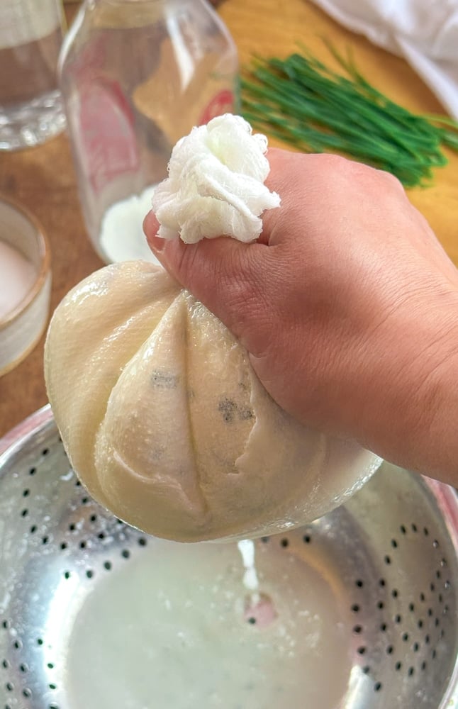 Hands squeezing a cheesecloth bundle of herbed cheese curds over a colander to drain the whey